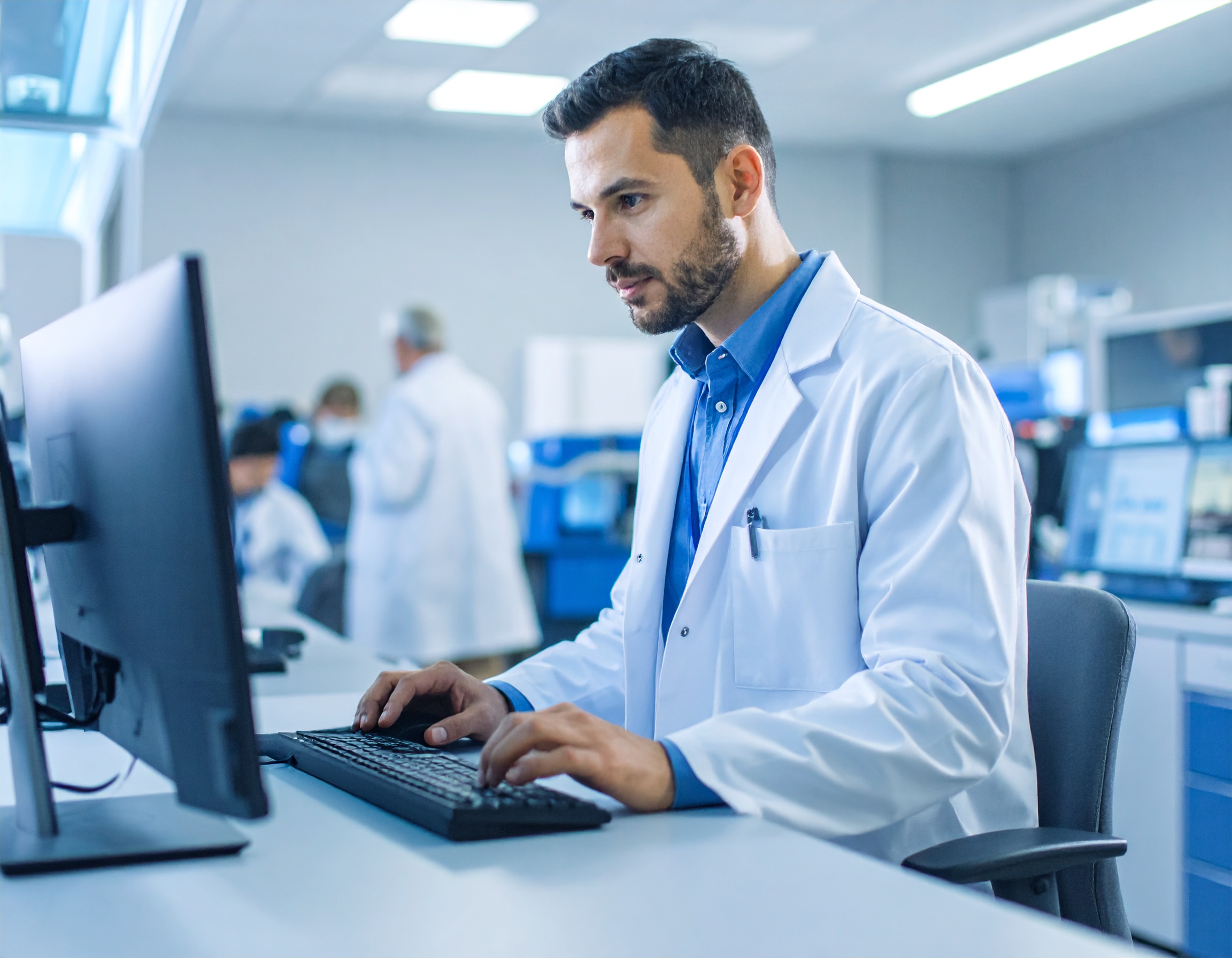 Scientist in a white lab coat typing on a keyboard at a computer in a laboratory.