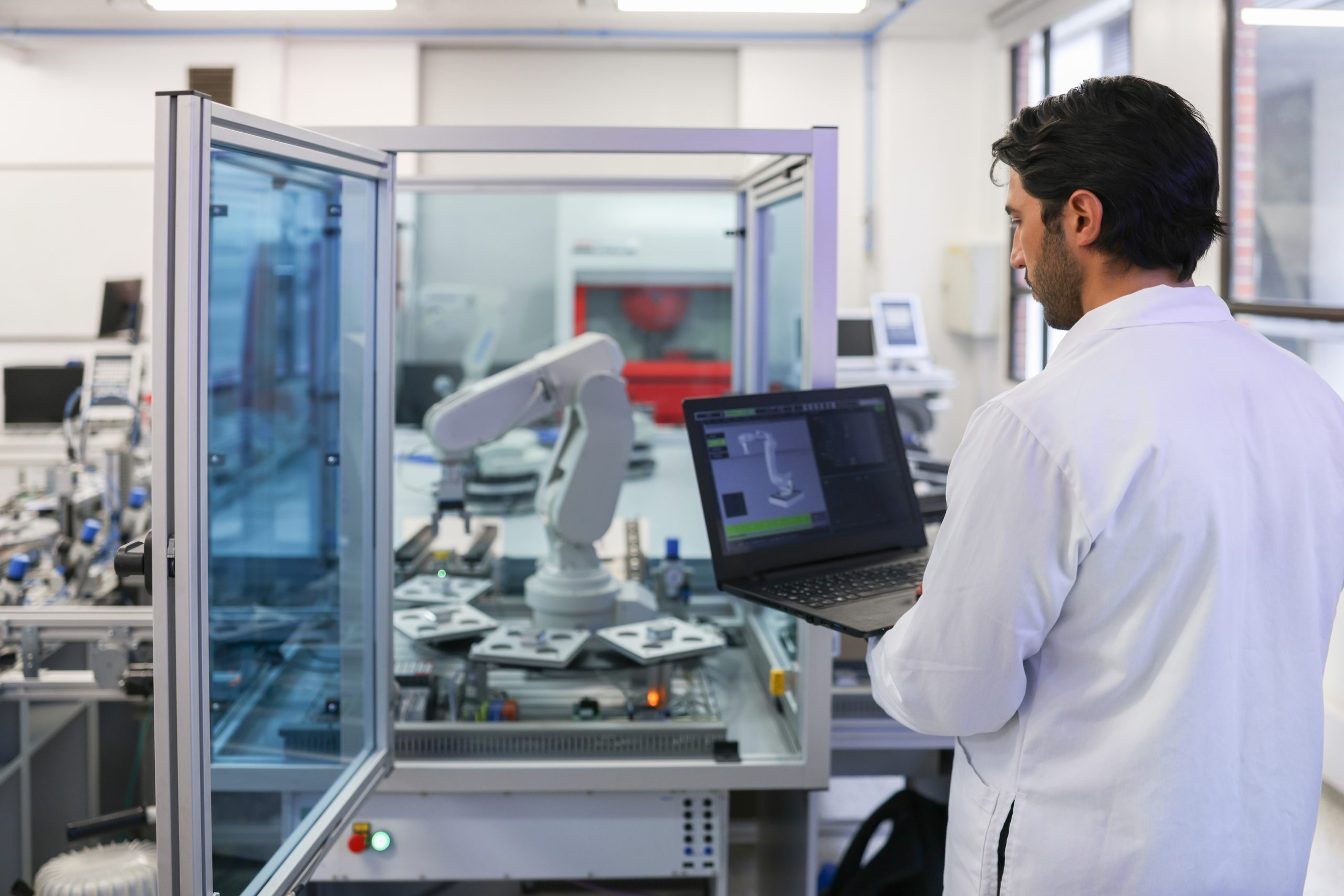 Lab technician in a white coat operates a computer near automated lab equipment inside a glass-walled enclosure in a cleanroom setting.