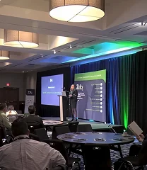 Presenter at a conference speaking from a podium on a stage with screens and blue/green lighting.