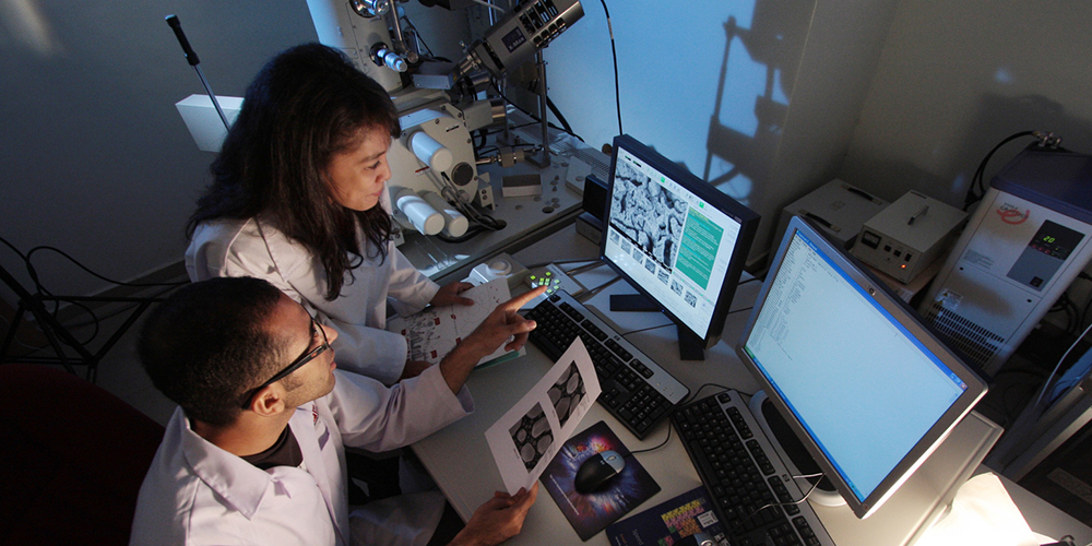 Two researchers in lab coats examine printed images at a workstation with microscopes and dual computer screens in a science lab.